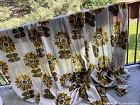Curtain fabric draped over balcony railing showing retro large floral sunflower pattern in olive and mustard tones on off-white background.