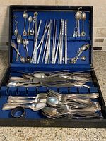 Photo showing opened wooden display box containing assorted vintage flatware pieces organized in felt compartments. Top lid holds collectible spoons with decorative bowls and handles.