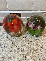 Two round glass paperweights, one with large red flower and the other with purple flowers and butterflies, placed on a granite countertop.