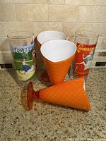 Set of three orange plastic waffle cone style sundae cups and two vintage highball glasses with colorful vintage graphics on a kitchen counter.