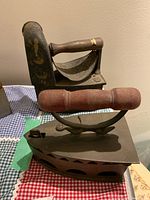 Two antique irons positioned side by side on a checkered and plain cloth, showing the painted decorative iron further back and the ventilation-hole iron in front.