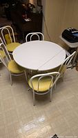 White drop leaf round breakfast table with 6 white metal frame yellow upholstered seats chairs in a kitchen area.