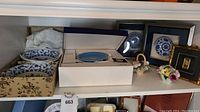 Wide view of shelf with boxed blue and white china plates and bowls, framed gold artwork, and ceramic flower arrangements.