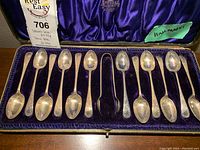 Full view of 12 silver teaspoons and one pair of silver tongs arranged in a blue velvet-lined case.