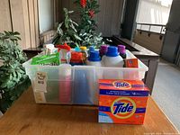 Wide view showing the lot of various laundry and cleaning supplies contained in a white bin on a wooden table.