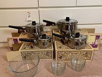 Full lot showing four stainless steel sauce pans with lids and black handles, arranged on original packaging boxes, with several clear ribbed glass bowls in front
