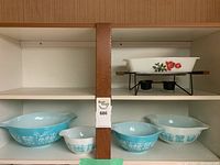 Shelf view showing four Pyrex mixing bowls on lower shelf and ceramic chafing dish on metal stand on upper shelf