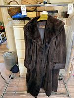 Front and partial side view of a long dark brown fur coat on a Siberian hanger, showing wide collar and buttoned cuff detail.