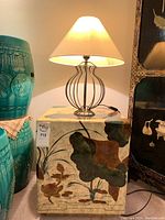 Front and side views of the decorative square side table with floral and leaf painting, next to a teal ceramic garden stool; topped with a black metal base lamp with cream conical shade.