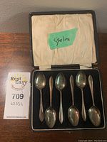 View of all seven sterling silver spoons inside the black lined case with satin lid interior and green paper label marked 'Sterling'. The spoons have decorative handles with a beaded or etched pattern.