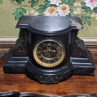 Front view of heavy antique eight day black mantel clock with gold Roman numerals and visible clockwork.
