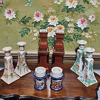 Front view of all eight candlestick holders arranged in two rows: three pairs of floral and bird ceramic/porcelain holders and one pair of red ceramic holders.