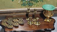 Set of brass accordion trivet, two pairs of brass candlestick holders, and a Trenton pottery pedestal vase/urn on a wooden surface