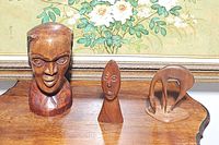 Photo showing all three carved wooden pieces: a large male head bust, smaller abstract mask figurine, and stylized deer statue on a wooden table.