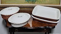 Four white ceramic casserole dishes, two rectangular each in a woven basket (one with lid, one without), and two oval covered casseroles (one with basket) arranged on a dark wood low table.
