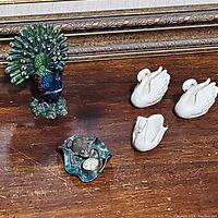 Five ceramic figurines displayed on a wooden surface placed before a framed painting: three white swans, one blue-green peacock with detailed feathers and pink tips, and one green frog with a watch.