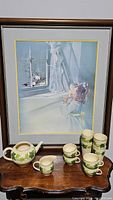 Franciscan Ivy china teapot, small pitcher, four teacups and four coffee mugs arranged on a wooden surface in front of framed artwork.