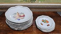 Stack of 8 larger Schumann dinner plates on left and 4 smaller Bavaria dessert plates on right showing scalloped edges and fruit designs.