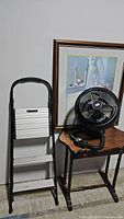 Photo showing the folded Cosco aluminum two-step stool with black handle and nearby black Bionaire fan on small table against a wall with framed picture.