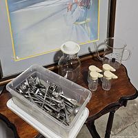 Wide view of all the items: plastic bin filled with stainless steel flatware, glass jar with white lid, four hinged salt and pepper shakers, a glass gravy separator, and a glass measuring cup on a small wooden table.