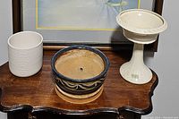 Three planters displayed on a wooden table: white ceramic pot, blue pottery planter with decorative scroll design and drainage hole, white pedestal planter by Gardenhouse