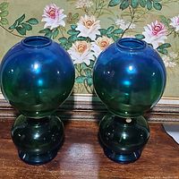 Pair of blue and green iridescent glass kerosene lamps side by side showing spherical shape and bases.