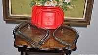 Three Pyrex glass baking dishes shown with red lids placed behind and beneath them on a small wooden table.