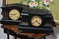 Two black mantel clocks displayed on wooden table, both with round faces and classical architectural designs.