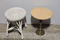 Two side tables side-by-side, showing white wicker table and brass pedestal table with plywood top in warehouse setting.