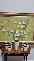 Full view of 10 stoneware cups and 1 jug arranged on wooden table under floral painting