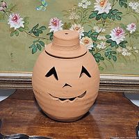 Front view of the large terracotta pumpkin pot showing smiling jack-o-lantern face with eyes, nose, and mouth cutouts, and lid on top.