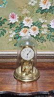 Full view of the vintage anniversary clock with brass base and clear glass dome, placed on wooden surface with floral painting background.