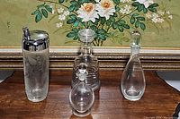 Front view showing four glass decanters and one cocktail shaker with chrome tops and stoppers, placed on a wooden surface against floral painting.