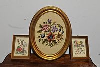 Three framed needlepoint pieces; one large oval floral needlepoint in gold frame, two smaller rectangular home-themed needlepoints in wooden frames.