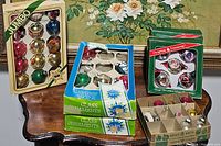 Boxes of multicolored shiny bulbs and vintage glass Christmas ornaments on a wooden table with floral wallpaper background.