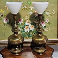 Pair of vintage brass Stiffel torchier lamps with white frosted glass shades on wood table in front of floral painting.
