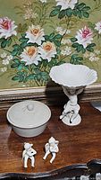Lot showing white ceramic pedestal cherub dish, pair of cherub figurines, and vintage crock on wooden surface