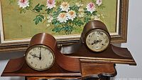 Two wooden mantel clocks side by side on a wooden surface in front of a floral painting, showing overall size and condition.