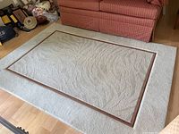Beige area rug laid on wood floor with textured wave design in center and brown border, partially under red striped couch.
