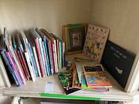 Bookshelf with children's books, some stacked and some standing upright, including visible titles The Velveteen Rabbit and This is Not My Hat, plus a framed teddy bear print leaning against the shelf wall.