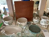 Collection of cream colored pottery pieces with botanical motifs displayed on glass shelf including cream and sugar bowls, leaf-shaped serving piece, additional bowls and green pottery plate