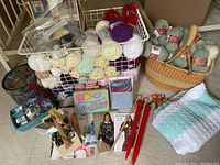 Overview of wool skeins in wire basket, wicker basket with green wool skeins and knitting needles, crocheted baby blanket, quilting placemat, and pattern booklets on the floor.