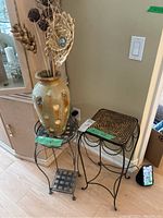 Photo of two black metal plant stands with decorative curved legs; left stand has two webbed metal shelves, right stand has woven rush top. A tall pottery vase with decorative motifs and dried floral arrangement is placed on left stand. The photo shows arrangement next to a mirror and light switch on wall.