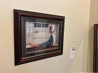 Framed painting of young girl sitting on porch, showing side profile with bonnet and blue dress, framed in dark wood with gold accents.