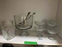 Glass trifle bowl with pedestal, two metal serving utensils, six clear stemmed dessert cups, and a glass water jug with fruit embossing on shelf.