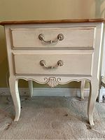 Front view of white washed nightstand showing two drawers, decorative handles, carved apron, and curved legs.