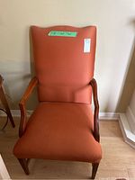 Front view of arm chair showing orange upholstery and wood arms and legs, with tape label on backrest showing measurements.