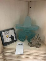 Three items on a shelf: a tall blue-green glass Inukshuk, a smaller layered soapstone Inukshuk, and a square Raku pottery tile with an Inuit figure and sun design.