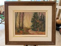 Framed oil painting on board depicting a forest scene with trees and a path, sitting on a wooden table.