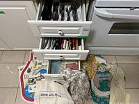 Kitchens drawers open showing neatly arranged flatware including butter knives, forks, and spoons. Several colorful aprons and tea towels laid out beneath the drawers.